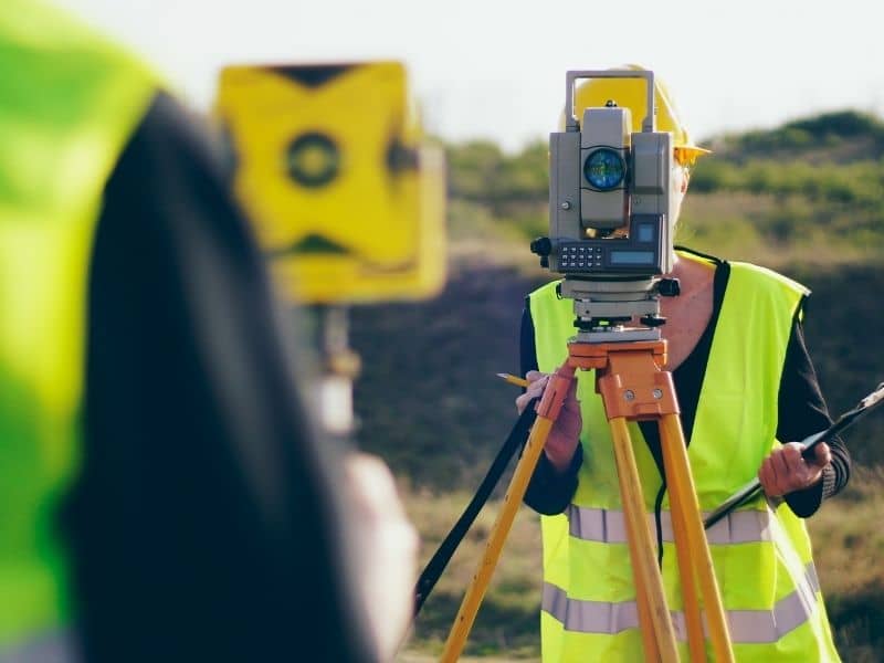 Boundary Survey Shared Privacy Wall Replacement Miami Florida | Miami Land Surveying Surveyor in a neon vest operates a total station on a tripod at a construction site outside.