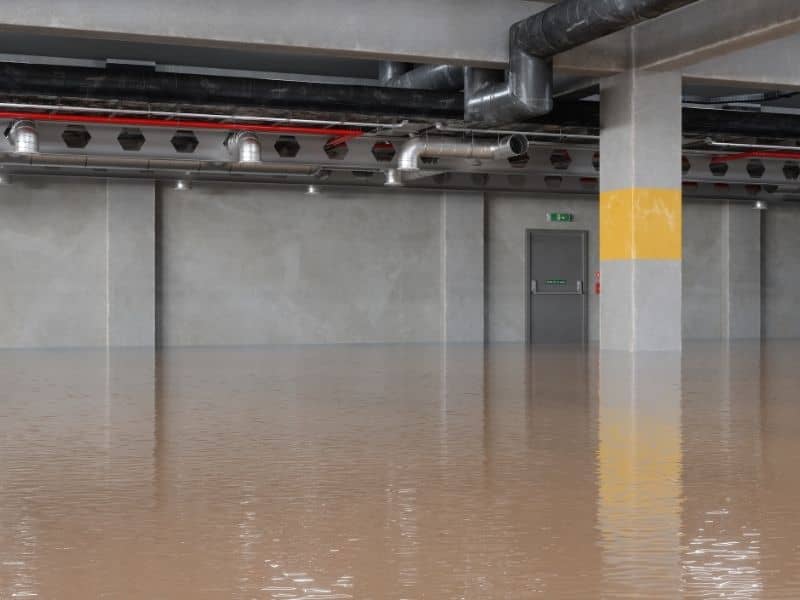 Basement with standing brown floodwater, exposed plumbing along the ceiling, and a gray door with an exit sign in the distance.