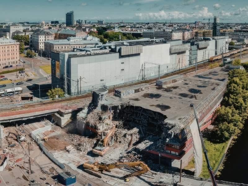 Aerial view of an urban demolition site with rubble and excavator near a railway viaduct, and a large modern building in the background.