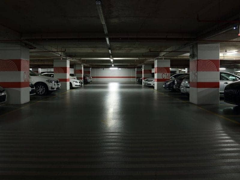 Underground parking garage with rows of parked cars along white pillars marked with red stripes; a bright center aisle leads to a distant exit/night lighting.